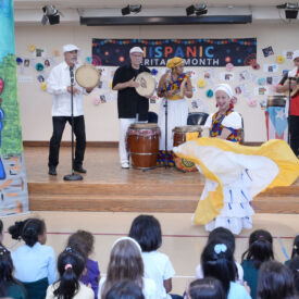 A woman in a brightly colored yellow skirt dances in front of a band. The band consists of two men playing drums, one man playing a guitar and one woman playing a handheld instrument.