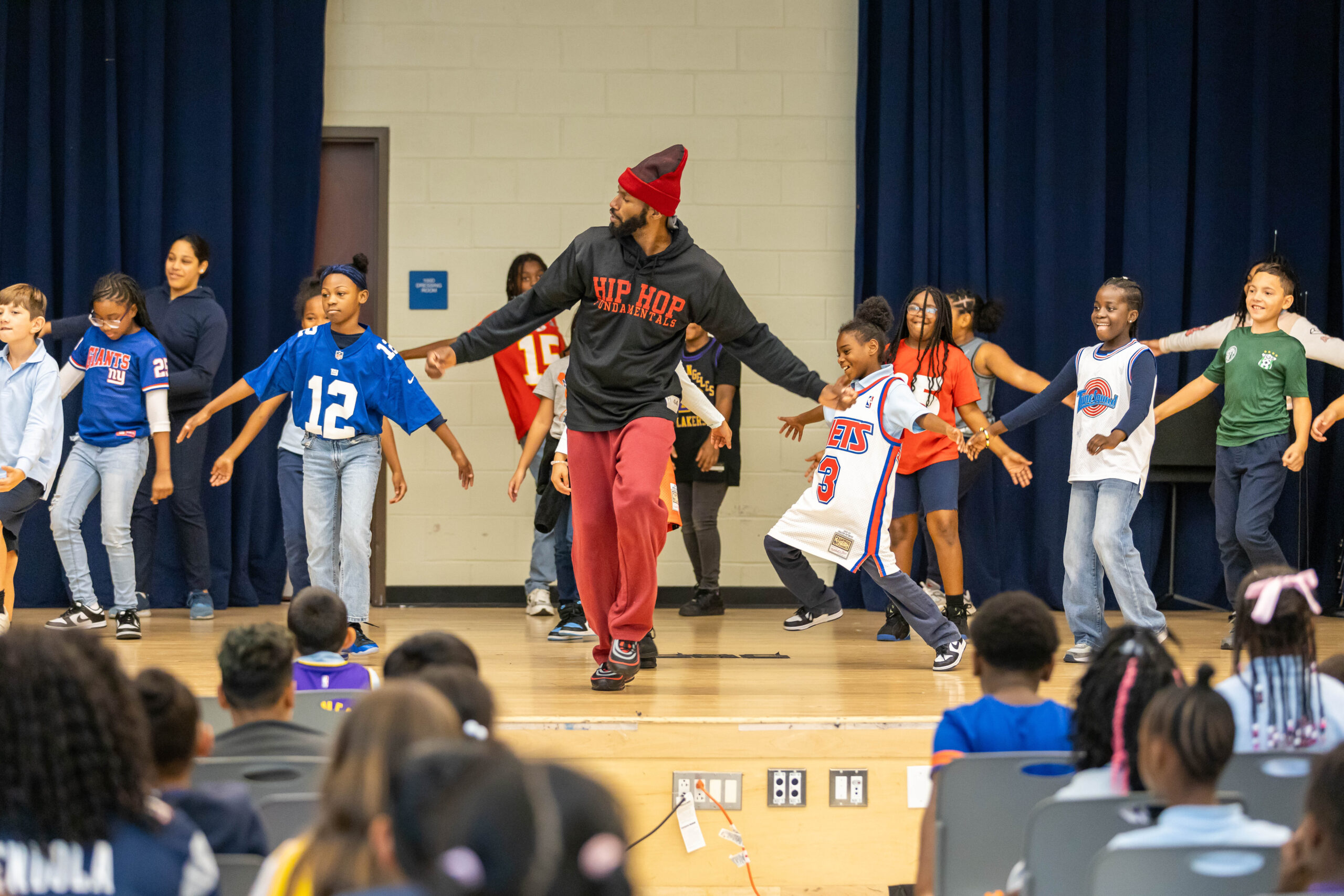 A Black man in a black hoodie and a red hat in the center of a group of children. His arms are out like he is breakdancing.