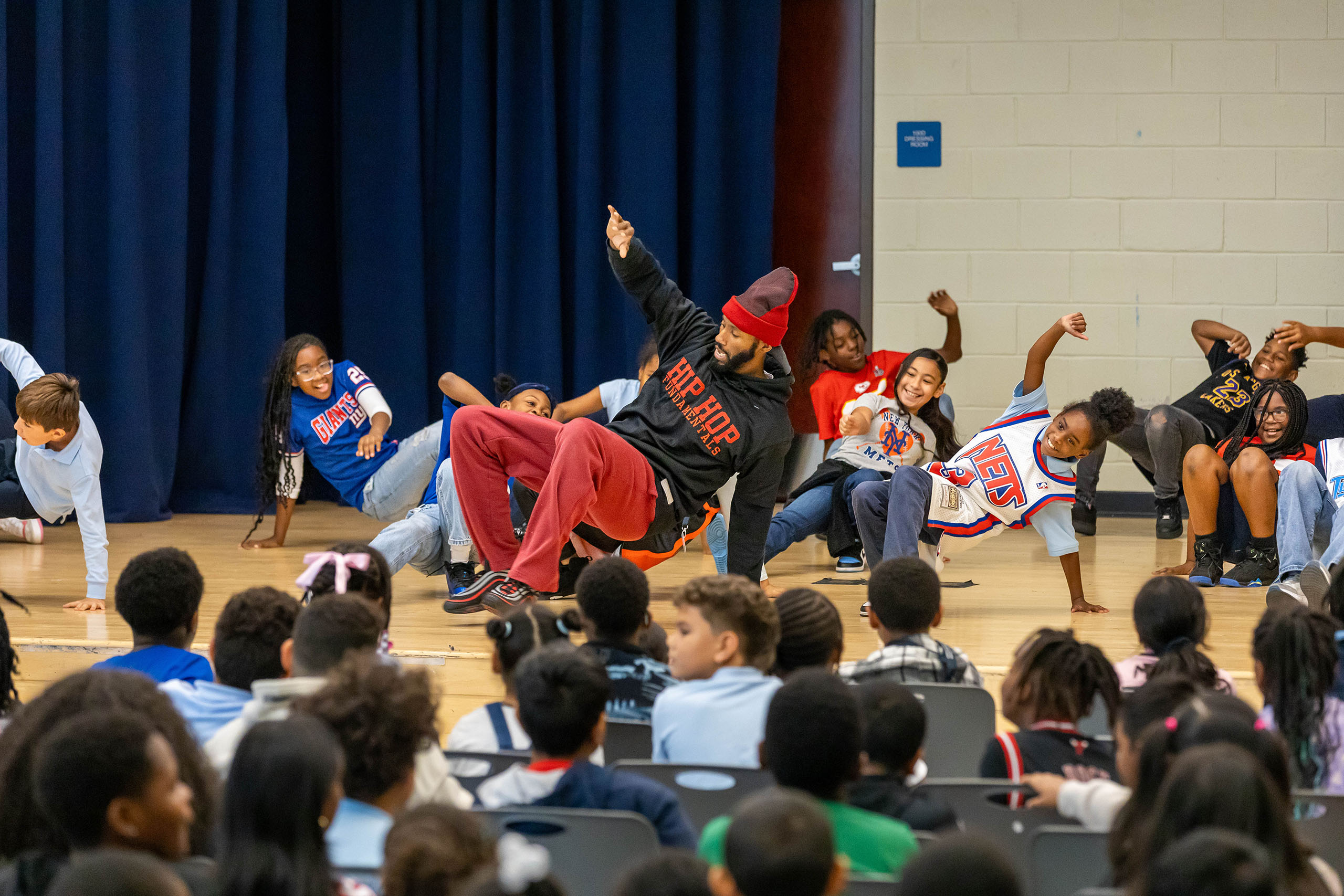 A Black man with a black hoodie and red pants breakdancing at a school with children behind him.