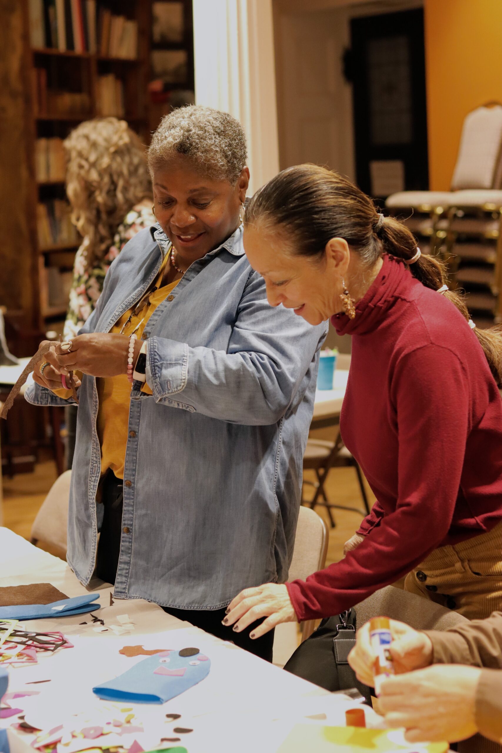 Woman with short grey hair cutting brown fabric on the right side with woman with long brown hair in a bubble ponytail style in red turtleneck looking down at table. 