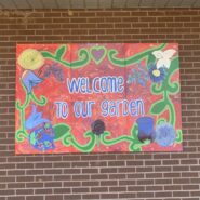 a large mural hanging on a brick wall that says "welcome to our garden" and has a border of flowers.