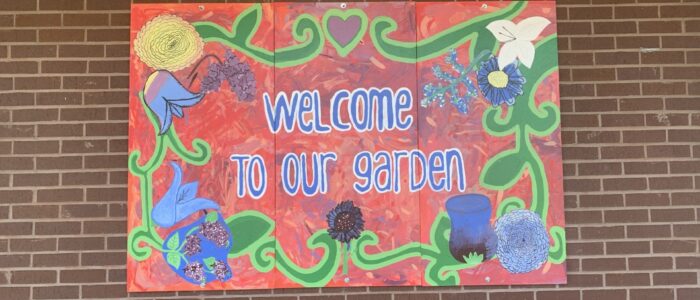 a large mural hanging on a brick wall that says "welcome to our garden" and has a border of flowers.