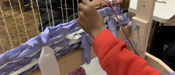 A student's hand using a giant loom.