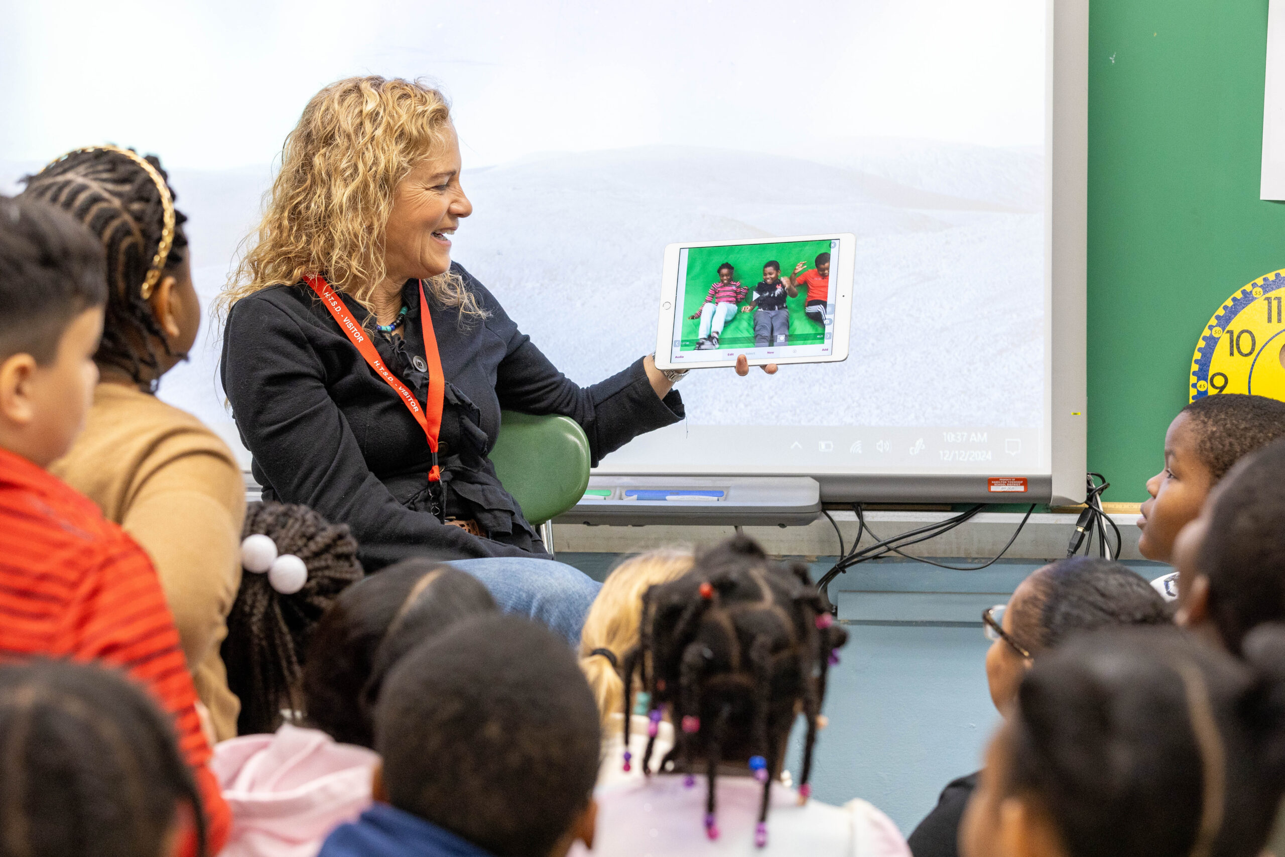 Alice Leon, a teaching artist, with blond hair, holds up a screen that shows students dancing with a green background. The students in the classroom are watching her attentively.