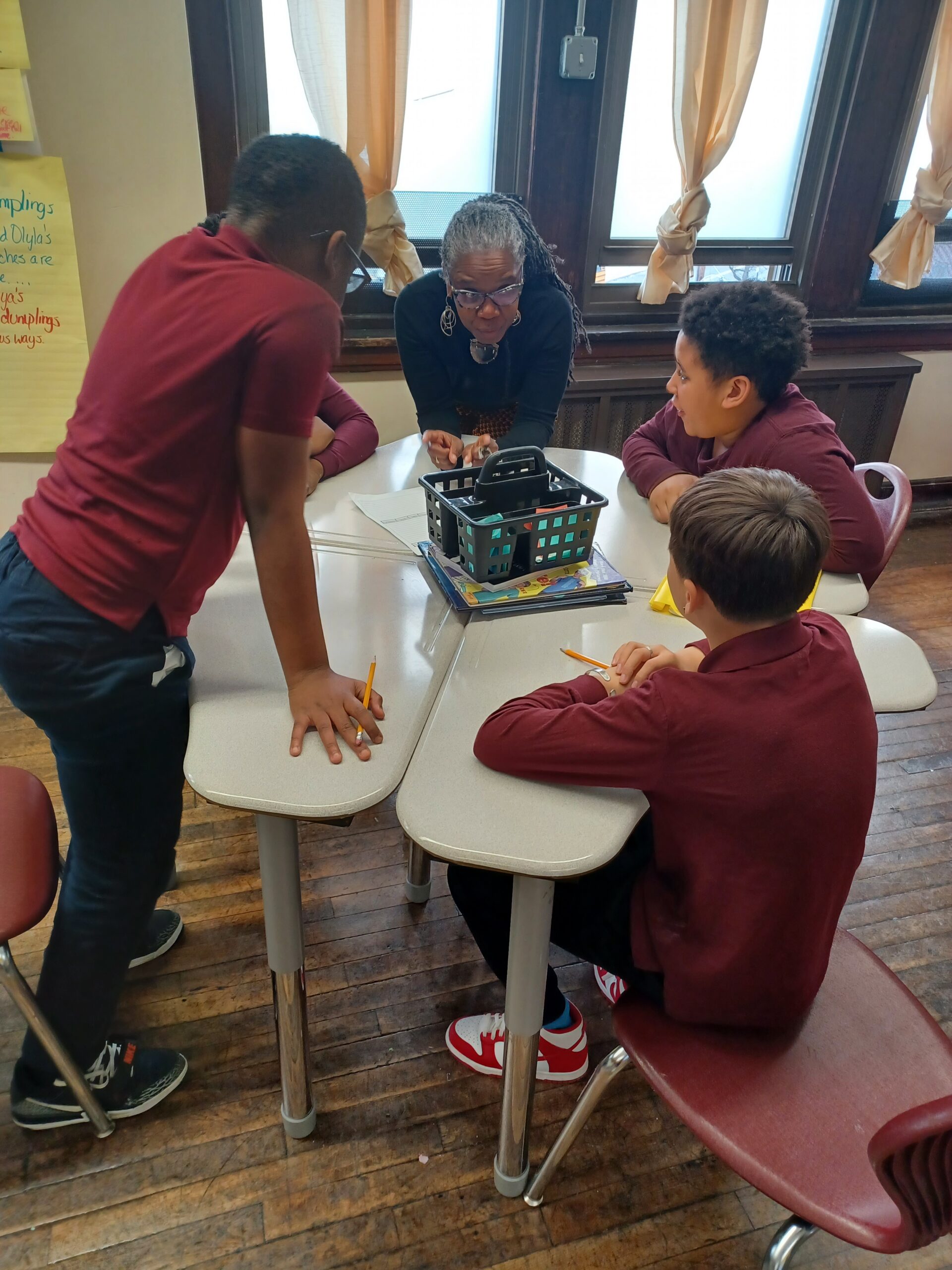 Teacher leaning over a table, guiding four students as they work on papers and supplies in a classroom. (Group learning scene)